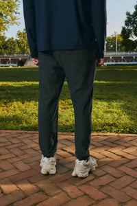 A person standing outdoors on a brick path in a park, wearing dark charcoal gray jogger pants, a navy long-sleeve top, and white sneakers.