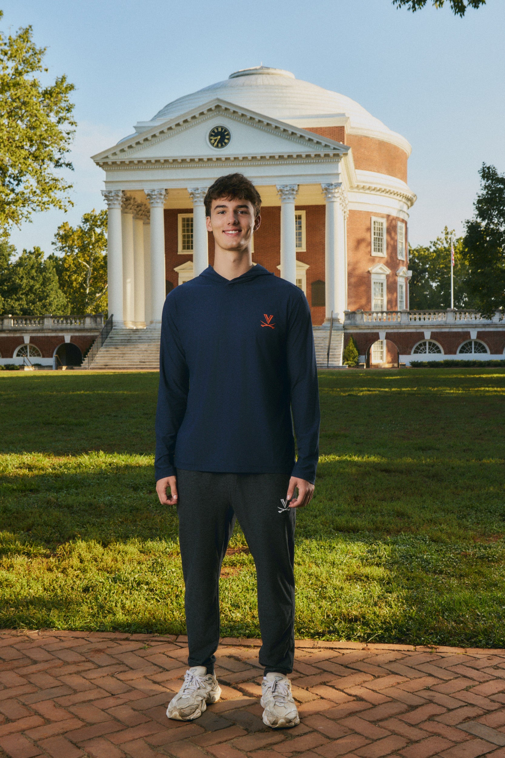 A young man stands outdoors in front of a large classical brick building on a grassy lawn, wearing a navy long-sleeve hoodie with a small red logo on the left chest, dark pants, and white sneakers.
