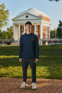 A young man stands outdoors in front of a large classical brick building on a grassy lawn, wearing a navy long-sleeve hoodie with a small red logo on the left chest, dark pants, and white sneakers.