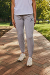 A person standing on a brick pathway in a park wearing heather gray jogger sweatpants with a navy Virginia Cavaliers logo on the upper left thigh, a white sleeveless top, and white sneakers.