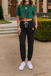 A woman modeling a dark green cropped t-shirt and black jogger sweatpants with a yellow 'BU' logo, standing outdoors on a concrete walkway in front of a brick building.
