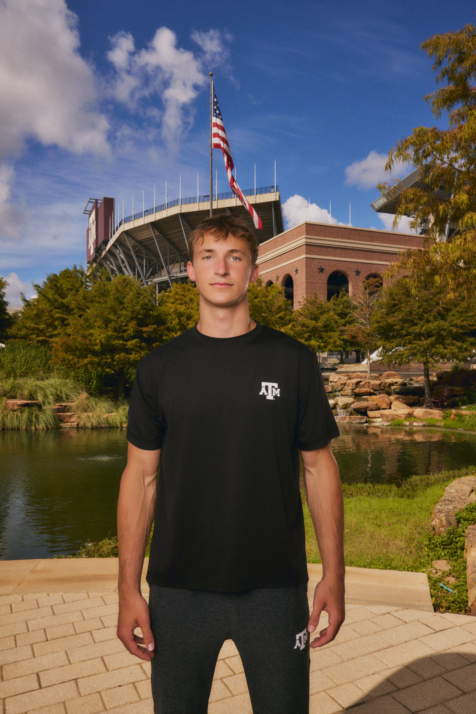 A man stands outdoors in front of a stadium and pond, wearing a black short-sleeve T-shirt with a small white Texas A&M logo on the left chest and dark gray sweatpants.