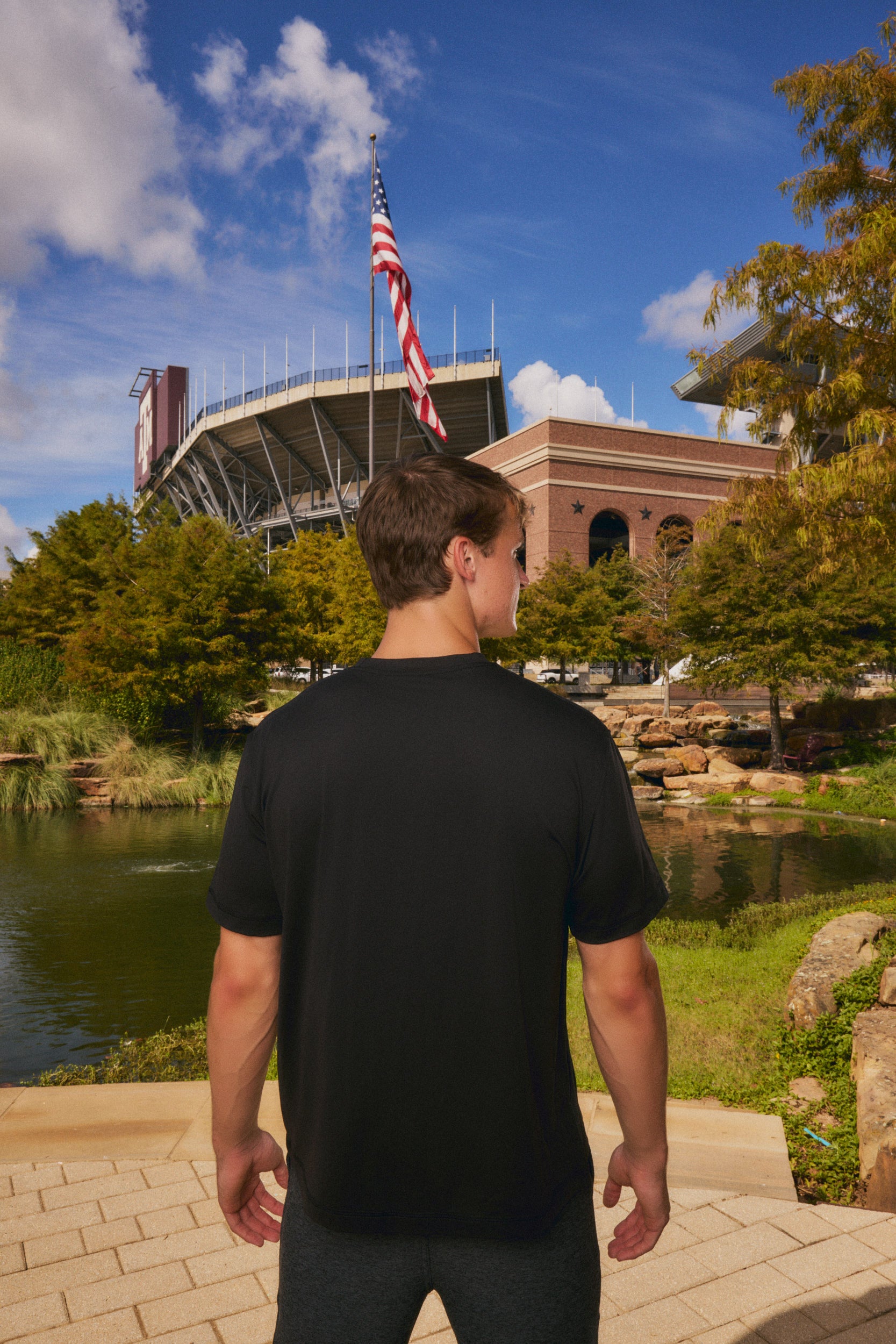 A man wearing a black short-sleeve athletic t-shirt stands with his back to the camera, looking towards a stadium and pond in an outdoor park setting under a blue sky.