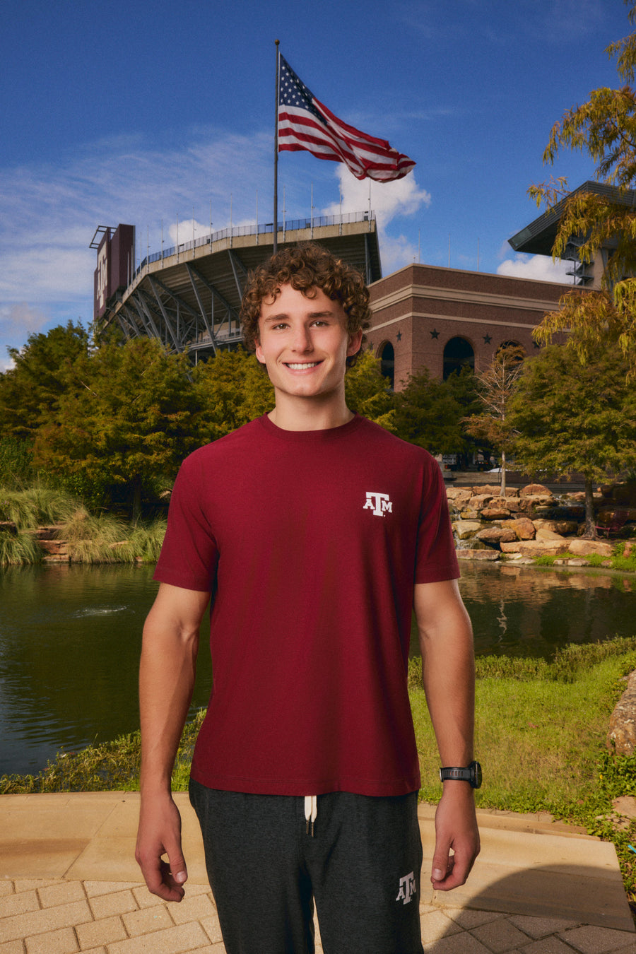 A smiling young man stands outdoors in front of a stadium and lake, wearing a burgundy short-sleeve T-shirt with a small white Texas A&M logo on the left chest and dark gray sweatpants.