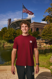 A smiling young man stands outdoors in front of a stadium and lake, wearing a burgundy short-sleeve T-shirt with a small white Texas A&M logo on the left chest and dark gray sweatpants.