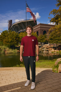 A smiling young man stands outdoors in front of a stadium and pond, wearing a maroon short-sleeve T-shirt with a small white Texas A&M logo on the left chest, black sweatpants, and white sneakers. An American flag is visible in the background.