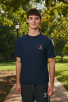 A young man standing on a brick path in a park, wearing a navy blue t-shirt with a small orange embroidered logo on the left chest and dark gray sweatpants.