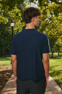 A man viewed from behind wearing a dark navy blue crewneck t-shirt and dark gray athletic shorts, standing on a brick pathway in a sunny park with green trees.