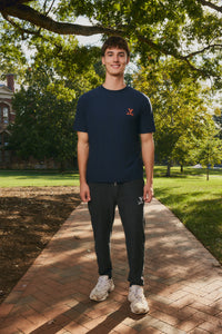 A man standing on a brick pathway in a park, wearing a navy blue t-shirt with a small orange logo on the left chest, dark gray sweatpants, and white sneakers.