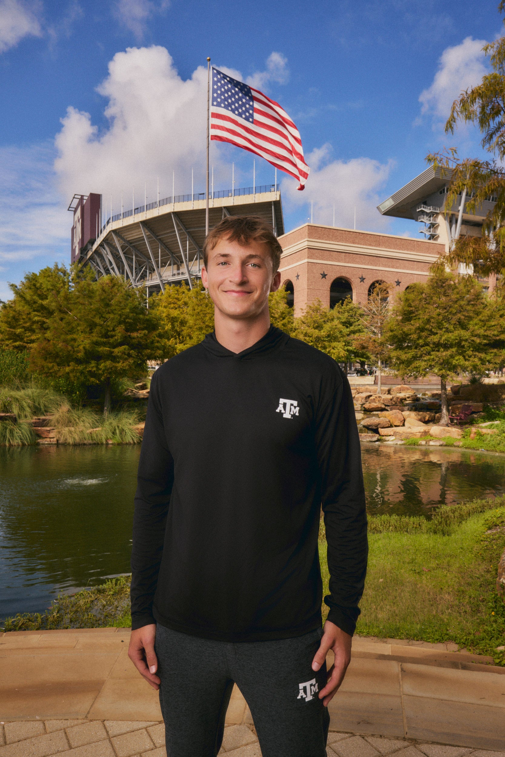 A man stands outdoors in front of a stadium and lake, wearing a black long-sleeve athletic pullover with a white Texas A&M logo on the chest and dark gray sweatpants with a matching logo on the thigh. An American flag flies behind him under a blue sky with clouds.