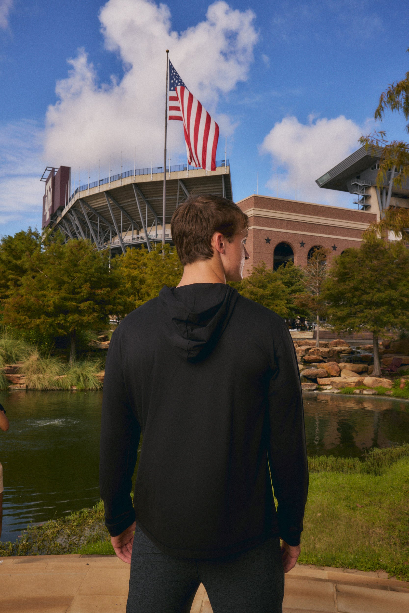 A man wearing a black hoodie stands with his back to the camera, looking towards a stadium with an American flag, set against a blue sky and green trees.