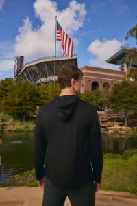 A man wearing a black hoodie stands with his back to the camera, looking towards a stadium with an American flag, set against a blue sky and green trees.
