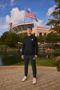 A man stands outdoors in front of a stadium and pond, wearing a black long-sleeve athletic top with a small white logo on the left chest, dark jogger pants, and white sneakers. An American flag is visible in the background.