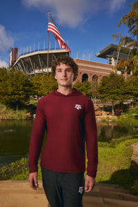 A young man stands outdoors in front of a stadium and pond, wearing a maroon long-sleeve hooded shirt with a white Texas A&M logo on the chest and dark sweatpants.