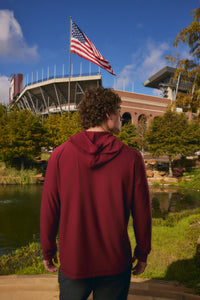 A man viewed from behind wearing a maroon hooded sweatshirt, standing outdoors in front of a stadium with an American flag, trees, and a pond.