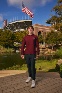 A young man stands outdoors in front of a stadium and pond, wearing a burgundy long-sleeve athletic hoodie with a small white logo on the left chest, black sweatpants, and white sneakers. An American flag flies above the stadium under a blue sky.