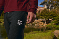 A person outdoors near a pond wearing dark heather gray sweatpants with a white Texas A&M 'A&M' logo on the thigh and a burgundy long-sleeve top. Trees, grass, rocks, and a building are visible in the background.