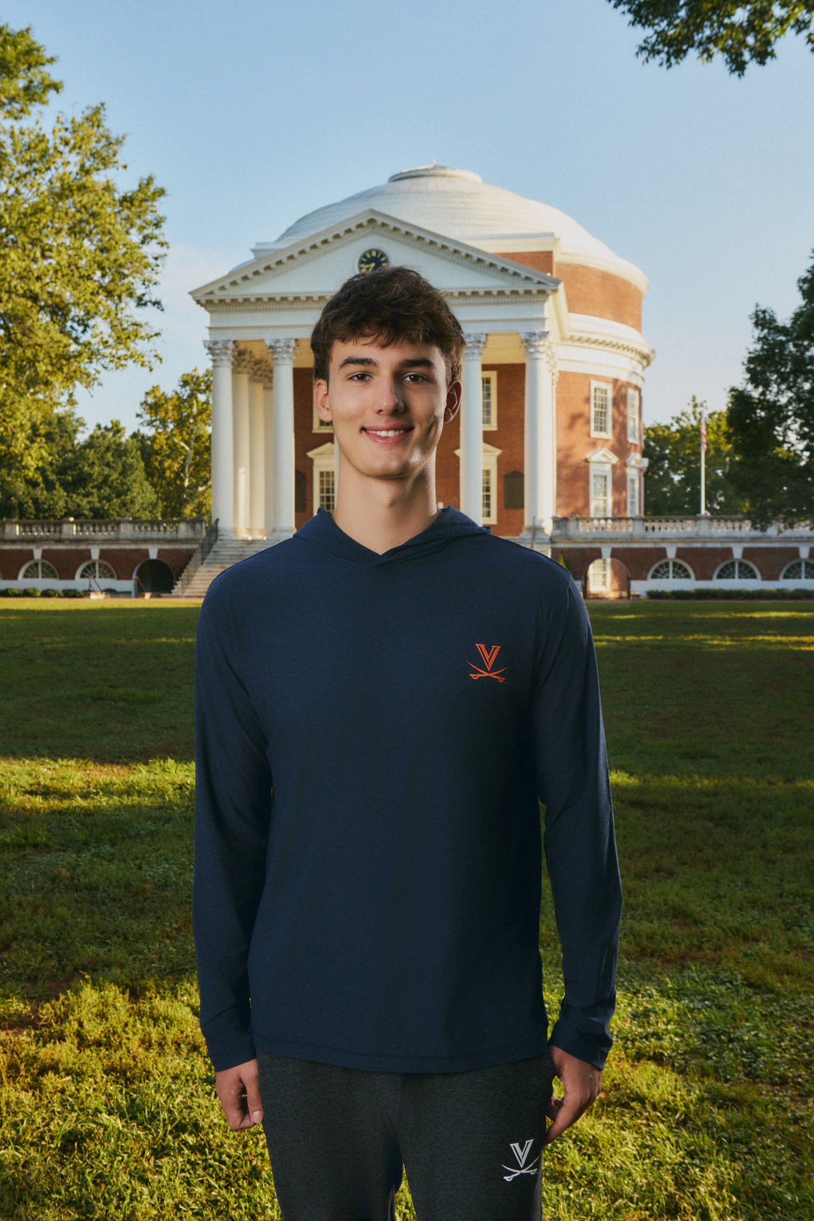 A young man stands outdoors in front of a large classical brick and white-domed building on a grassy lawn. He is wearing a navy blue long-sleeve hooded shirt with a small red logo on the left chest and dark gray sweatpants.