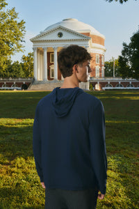 A man wearing a navy blue hooded sweatshirt stands on a grassy lawn with a historic domed brick building in the background, photographed from behind.