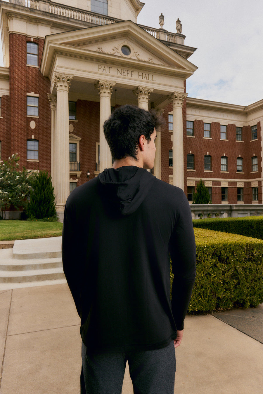 A man wearing a black long-sleeve athletic hoodie and dark gray sweatpants, photographed from behind as he stands in front of a large classical brick building with columns. The building has 'PAT NEFF HALL' engraved above the portico.