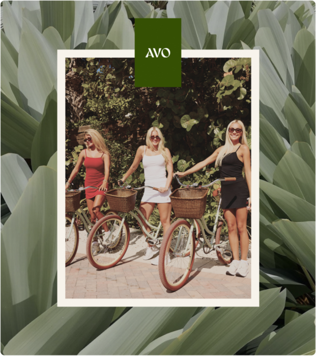 A square lifestyle photo with a white border and green AVO logo at the top, showing three women standing outdoors with vintage bicycles. The photo is centered over a background of large green leaves.