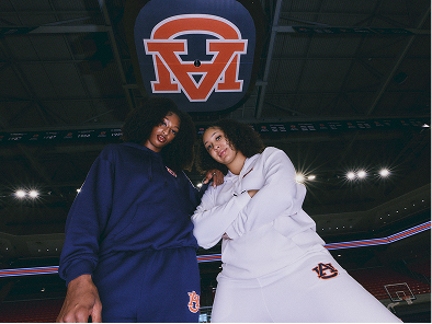 Two women posing in Auburn University athletic apparel inside a basketball arena. One wears a navy blue tracksuit and the other wears a white tracksuit, both featuring the Auburn 'AU' logo.