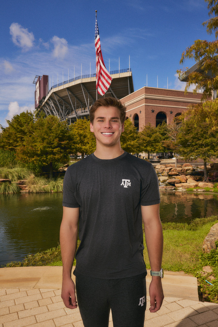 A smiling young man stands outdoors in front of a stadium and pond, wearing a dark charcoal gray athletic t-shirt with a white 'ATC' logo on the chest and matching dark athletic pants.