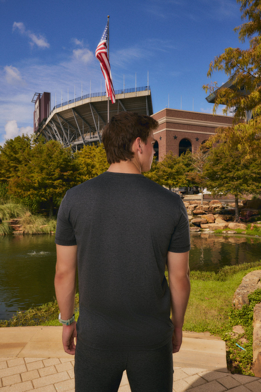 A man wearing a dark gray heathered t-shirt and matching shorts stands with his back to the camera, looking towards a stadium and pond in an outdoor setting.