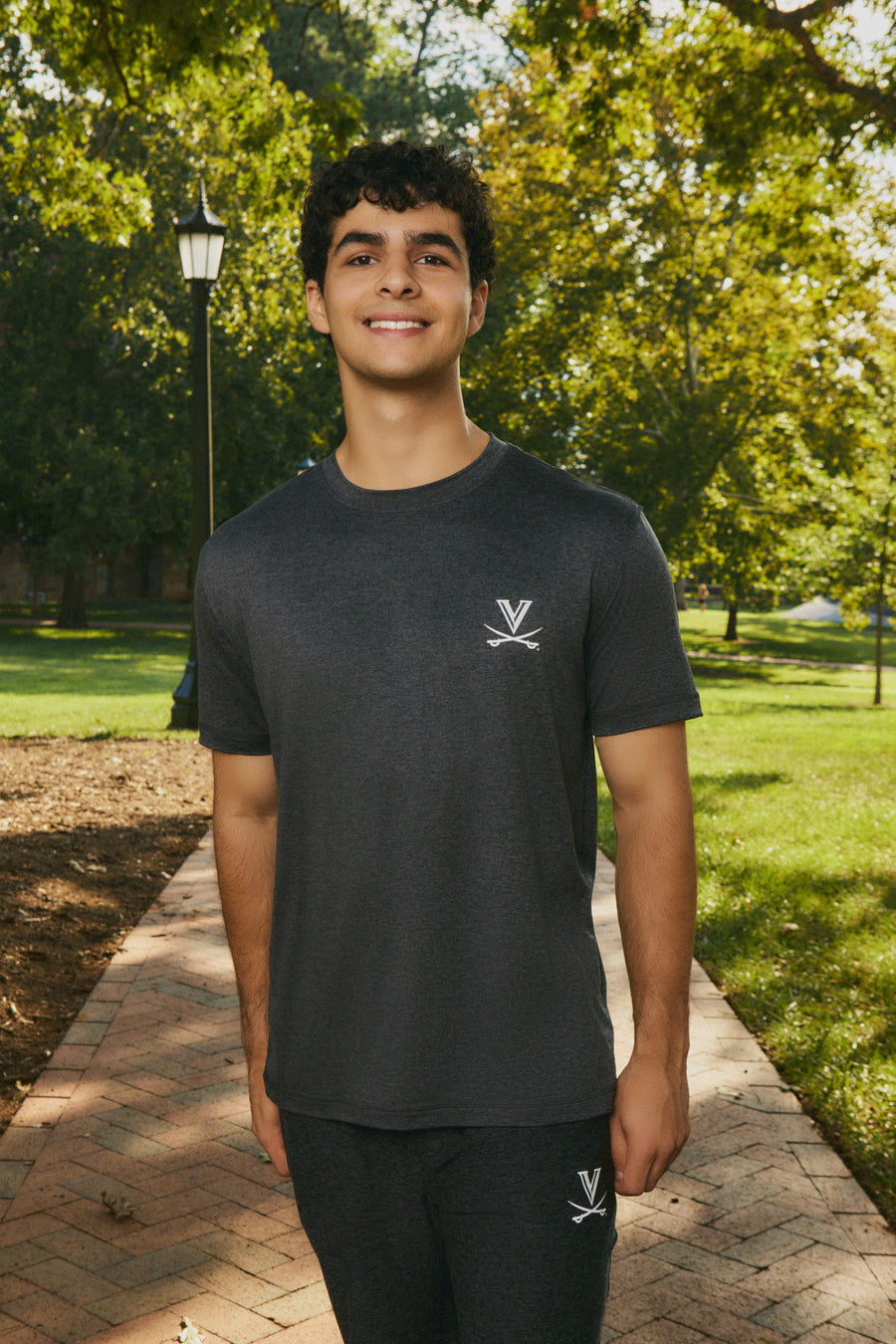 A young man smiling while standing on a brick pathway in a sunny park, wearing a dark charcoal gray short-sleeve t-shirt with a small white Virginia Cavaliers logo on the left chest and matching dark gray athletic pants.