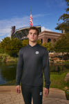 A man stands outdoors in front of a stadium and pond, wearing a dark gray long-sleeve athletic hoodie with a white Texas A&M logo on the chest and matching dark athletic pants.