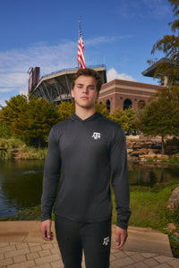 A man stands outdoors in front of a stadium and pond, wearing a dark gray long-sleeve athletic hoodie with a white Texas A&M logo on the chest and matching dark athletic pants.