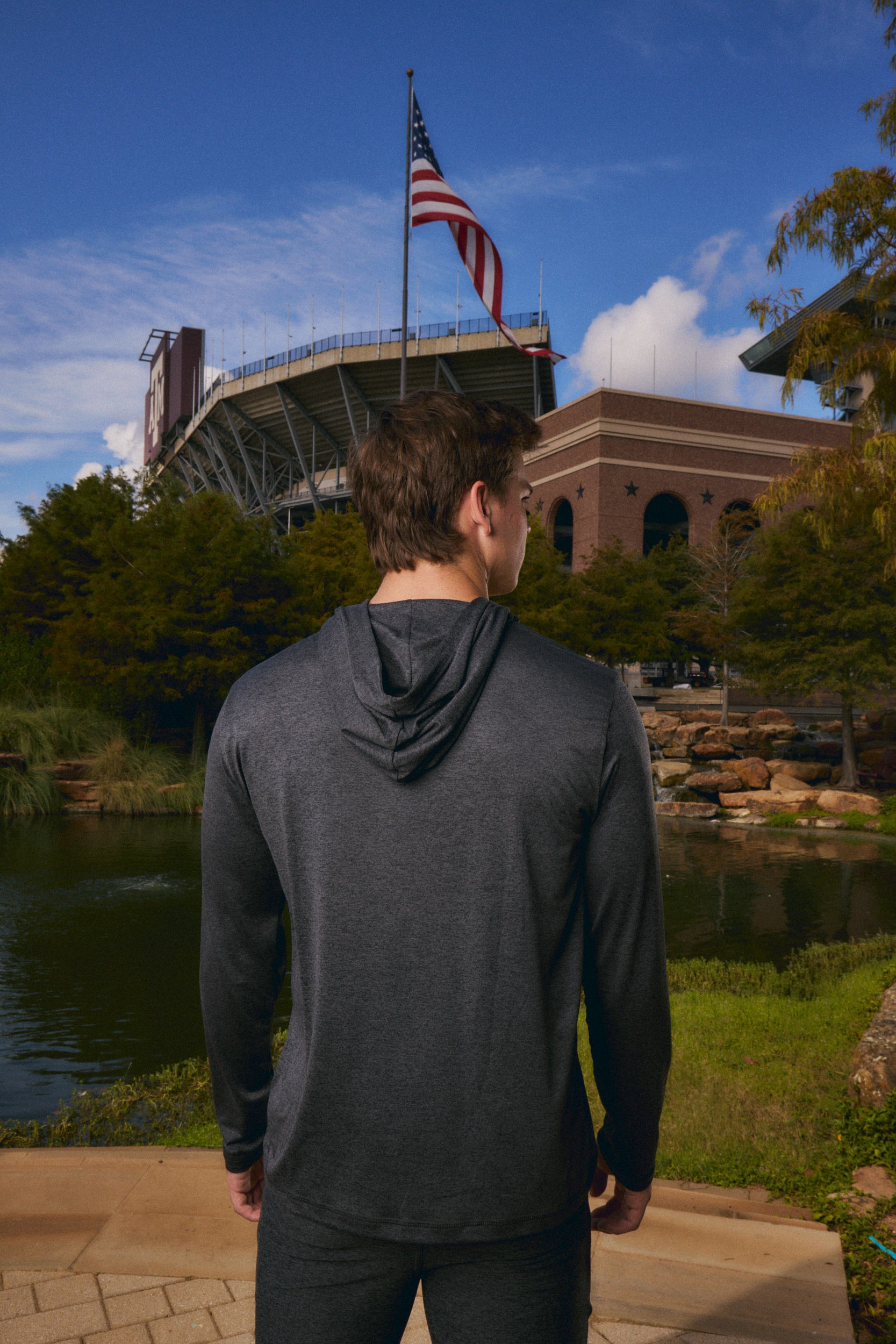 A man viewed from behind wearing a dark gray long-sleeve athletic hoodie and black pants, standing outdoors in front of a stadium and pond with an American flag in the background.