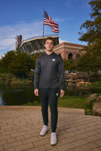 A man wearing a dark gray long-sleeve athletic hoodie and matching black track pants stands outdoors on a brick walkway in front of a stadium, with a pond, trees, and an American flag in the background.