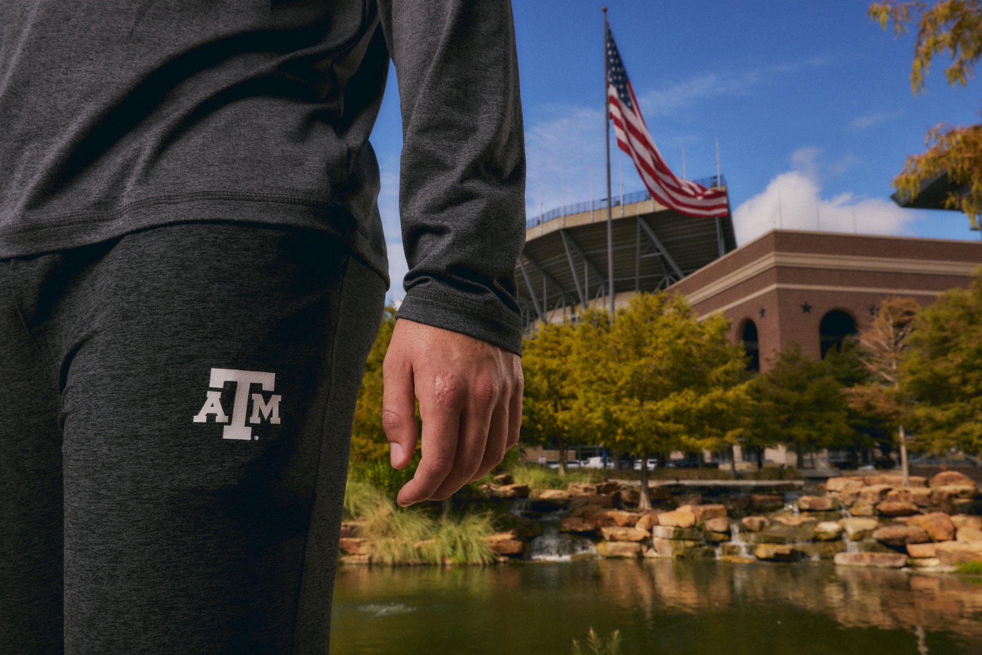A person wearing a dark gray long-sleeve shirt and matching sweatpants with a white Texas A&M logo, standing outdoors near a pond with stadium architecture and an American flag in the background.