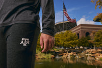 A person wearing a dark gray long-sleeve shirt and matching sweatpants with a white Texas A&M logo, standing outdoors near a pond with stadium architecture and an American flag in the background.