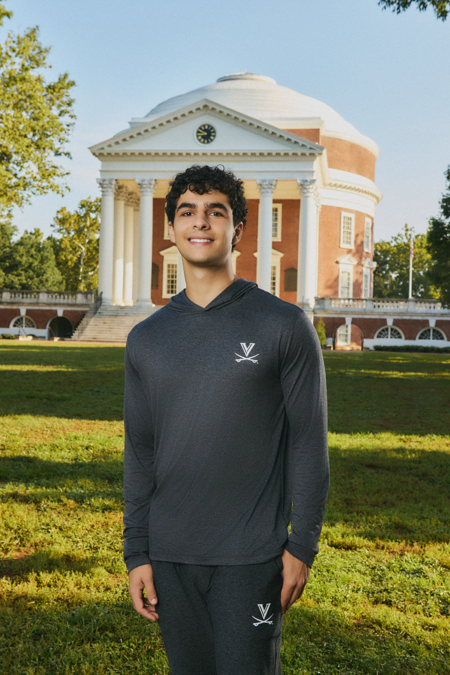 A young man wearing a dark gray long-sleeve athletic hoodie and matching sweatpants stands on a grassy lawn in front of a large classical brick and white-domed building. The hoodie has a small white logo on the left chest, and the sweatpants have a matching logo near the lower leg.