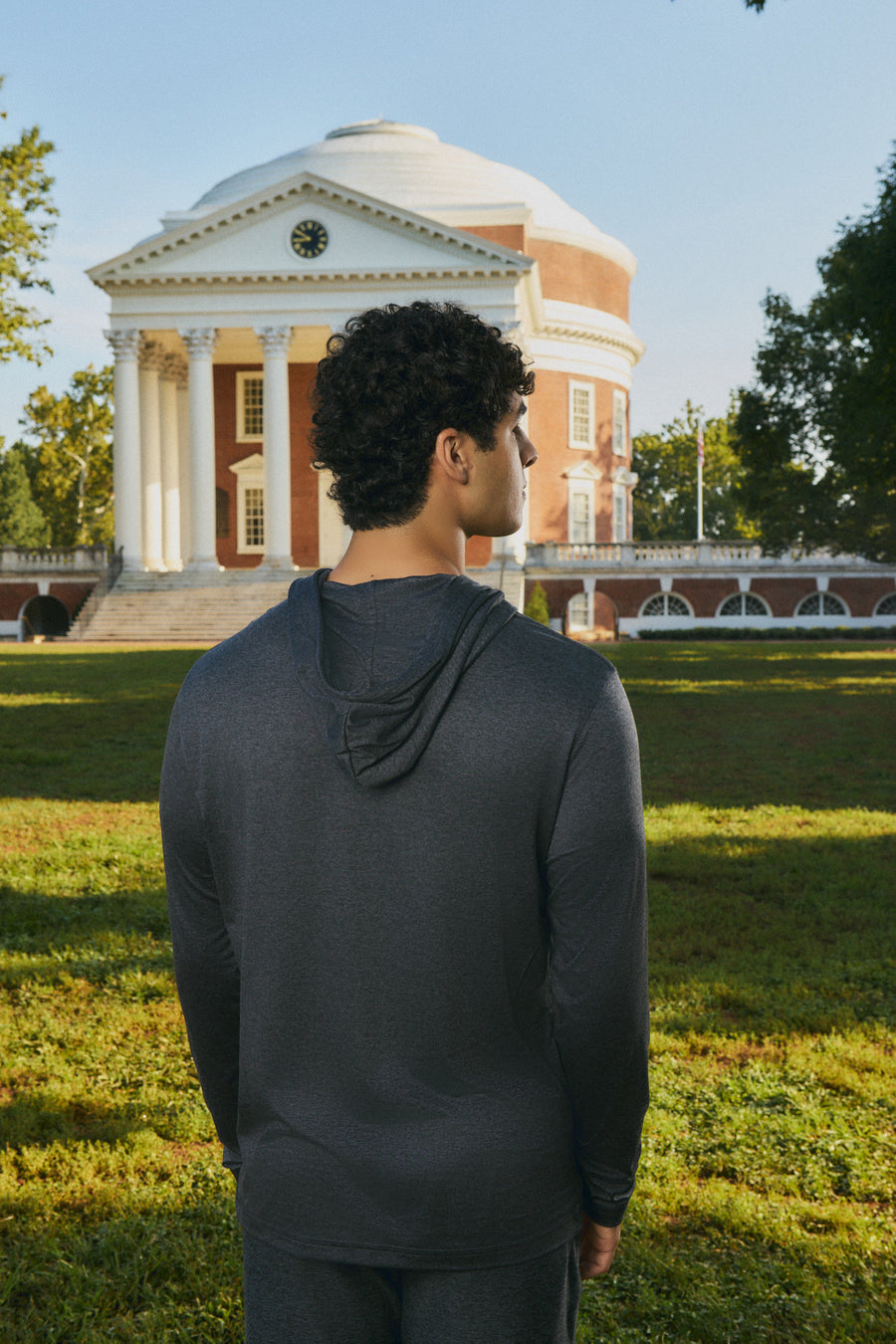 A man wearing a dark gray long-sleeve athletic hoodie stands with his back to the camera, looking toward a historic red-brick domed building on a sunny grassy lawn.