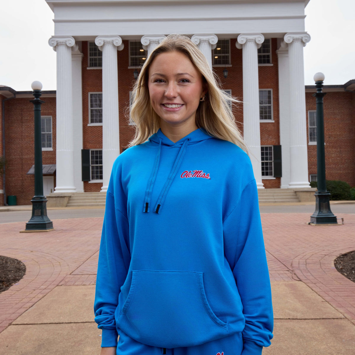 A smiling blonde woman stands outdoors in front of a brick building with white columns, wearing a bright blue pullover hoodie with 'Ollie's' embroidered in red on the chest.