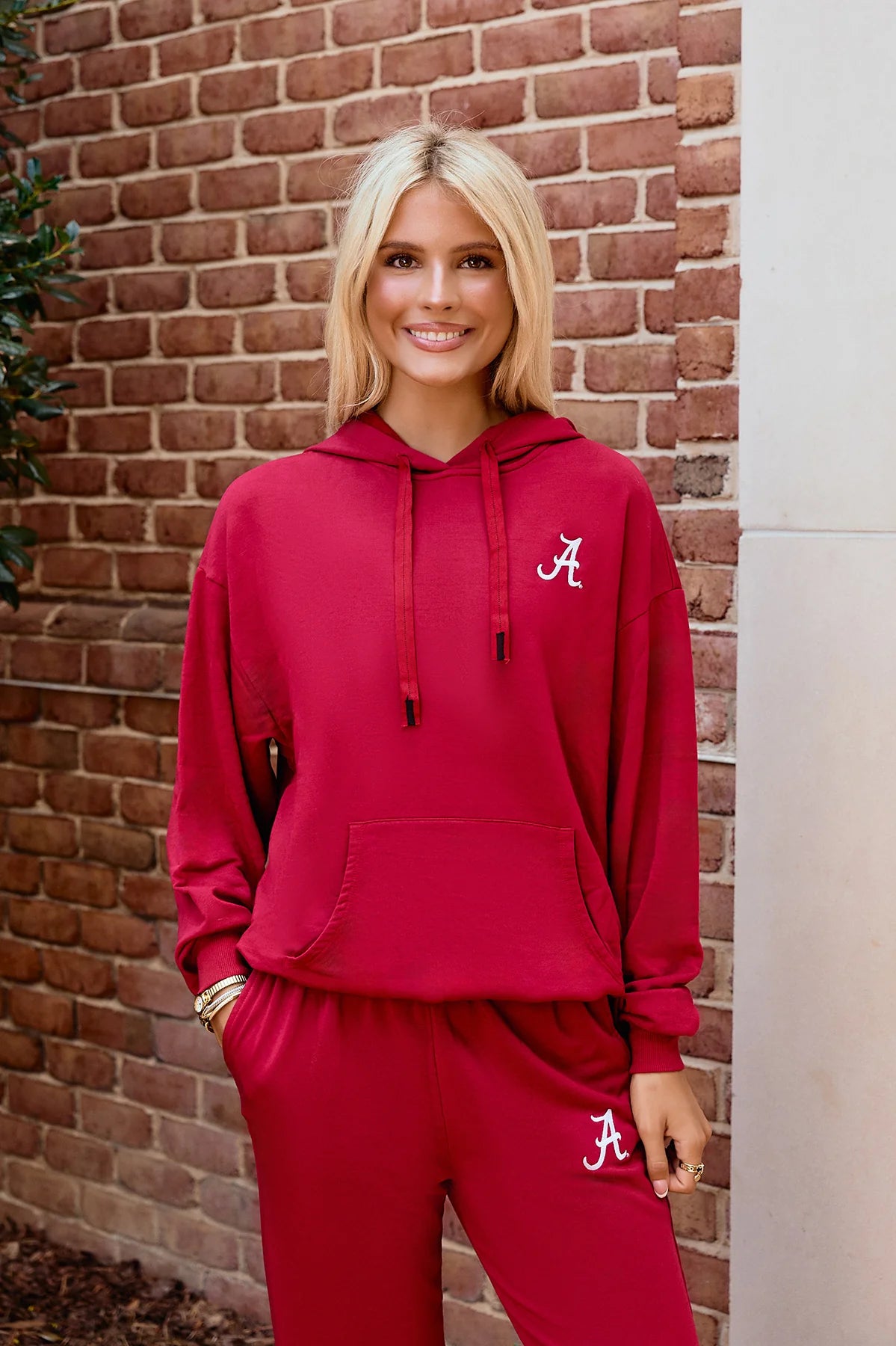 Woman wearing a crimson Alabama hoodie and matching sweatpants with white “A” logo embroidery, posing against a brick wall. The coordinated lounge set features a relaxed fit, front kangaroo pocket, and drawstring hood in a casual collegiate style.
