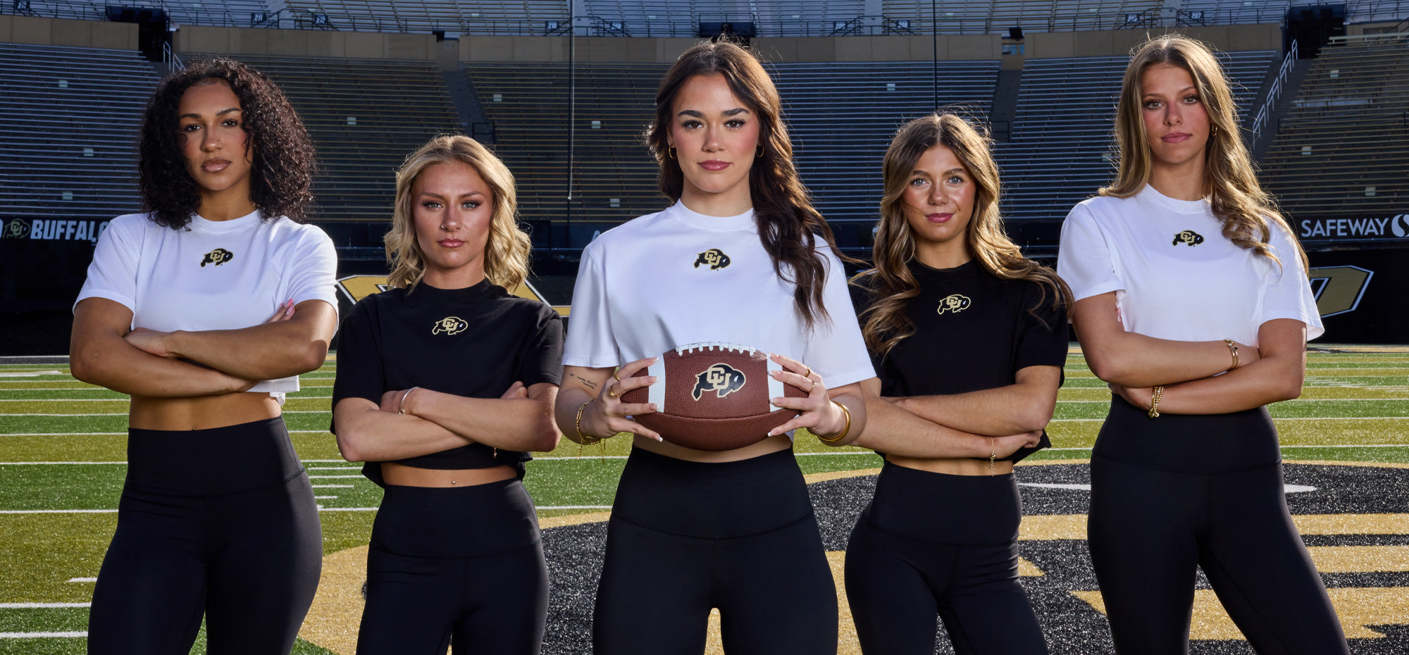 Five women standing on a football field wearing Colorado Buffaloes apparel, with the center woman holding a football