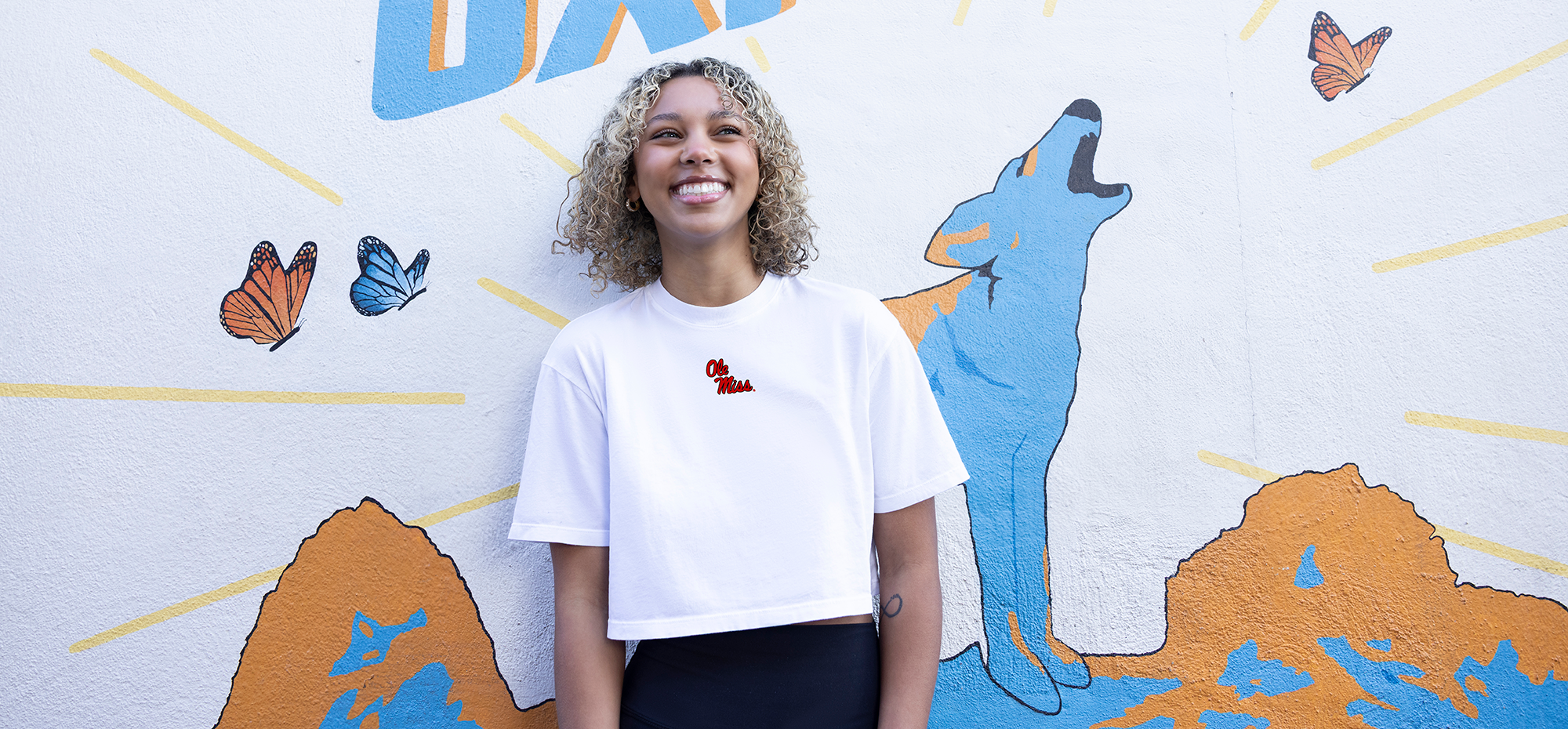 Woman wearing an Ole Miss shirt standing in front of a colorful mural with butterflies and a dog illustration