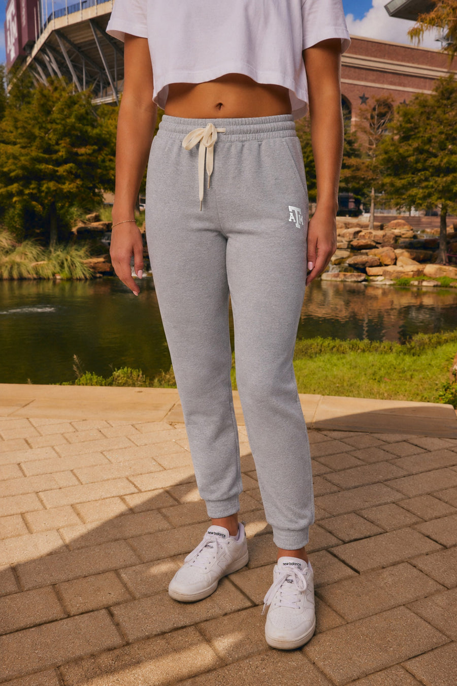 A person wearing light gray jogger sweatpants with a cream drawstring, shown from the waist down outdoors near a pond and brick walkway.